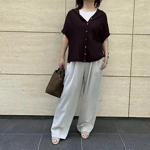 濃い赤茶色のボタンダウンシャツに白いタンクトップを合わせ、白いワイドパンツとスニーカーを着用した女性。右手に茶色のバッグを持ち、正面を向いている。背景はベージュのタイル壁。