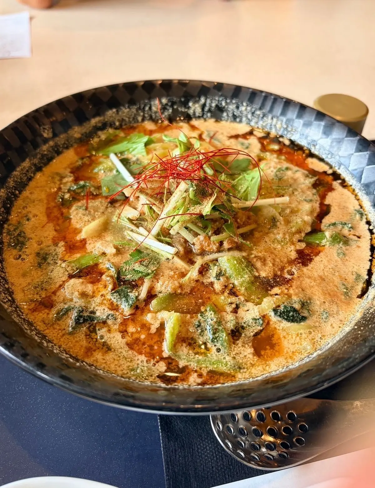 黒い鉄製の鍋に入った、オレンジ色のスープ。緑色の野菜と細切りになったネギ、赤い唐辛子が散らされている。