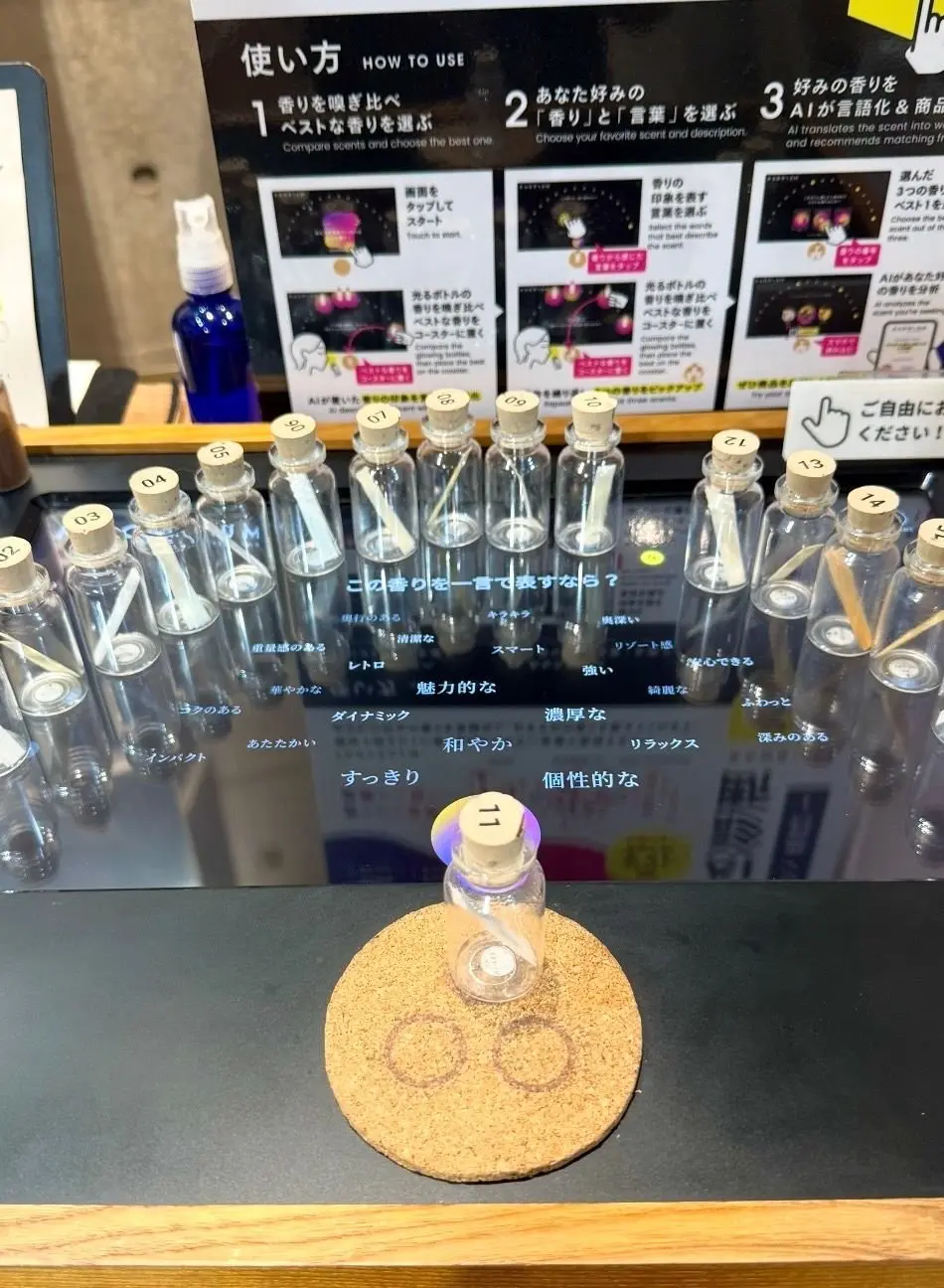 香水試香台。透明なガラス瓶が並び、それぞれに番号が振られている。前面には使用方法の説明書きと、香りの特徴を表す言葉が書かれたカードが置かれている。中央には紫色のディフューザーが置かれている。