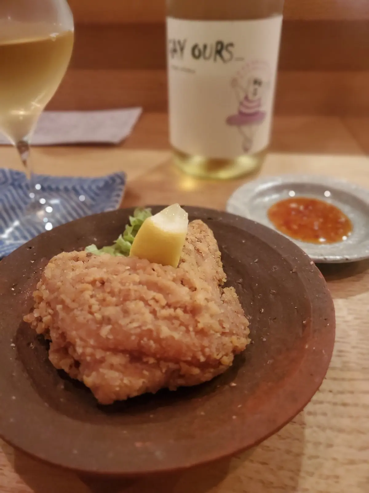 木製のテーブルに置かれた、揚げ鶏（唐揚げ）の盛り合わせ。粗めの陶器皿に盛り付けられ、レモンが添えられている。背景にはワインボトルとグラス、小皿が写る。