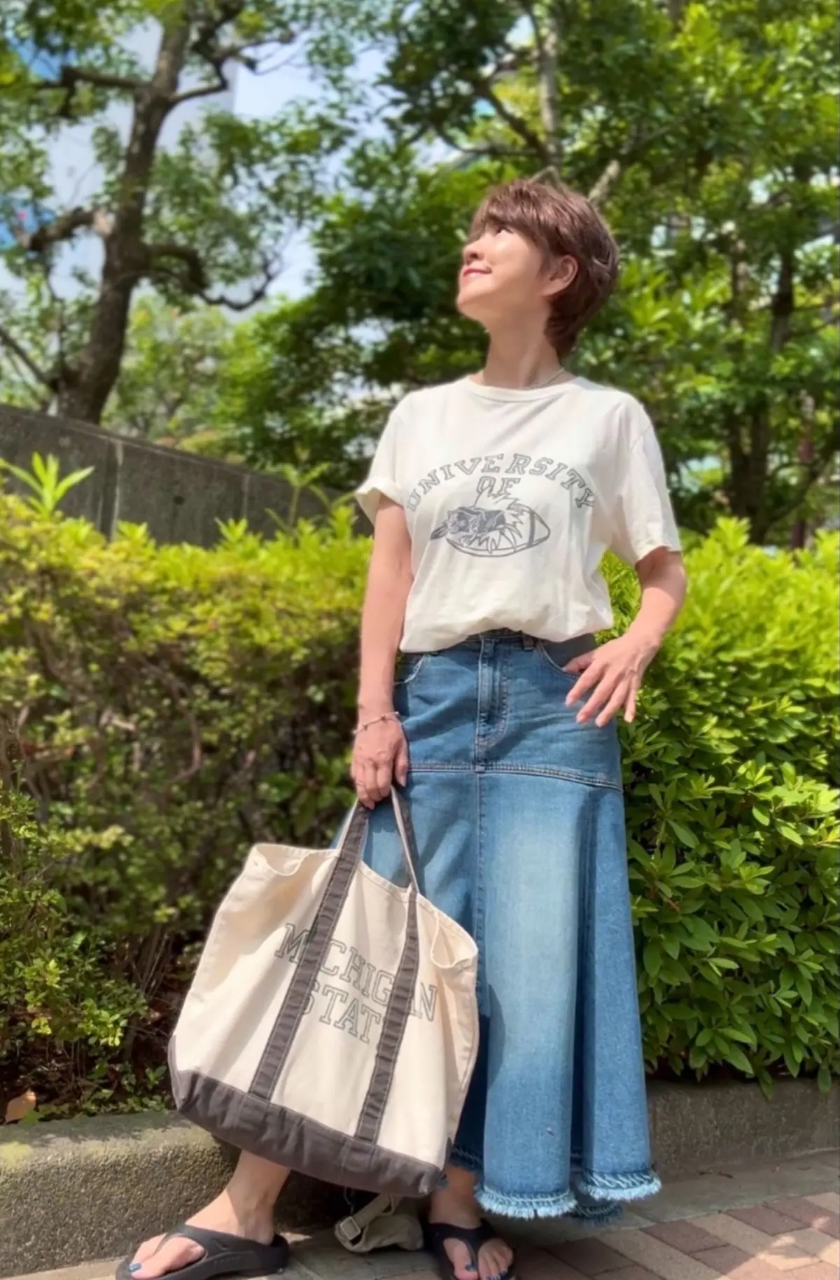 女性が白い大学ロゴ入りTシャツとデニスカジュアルスカートを着て、緑豊かな公園で晴天の下、ベージュのトートバッグを手に持って立っている。