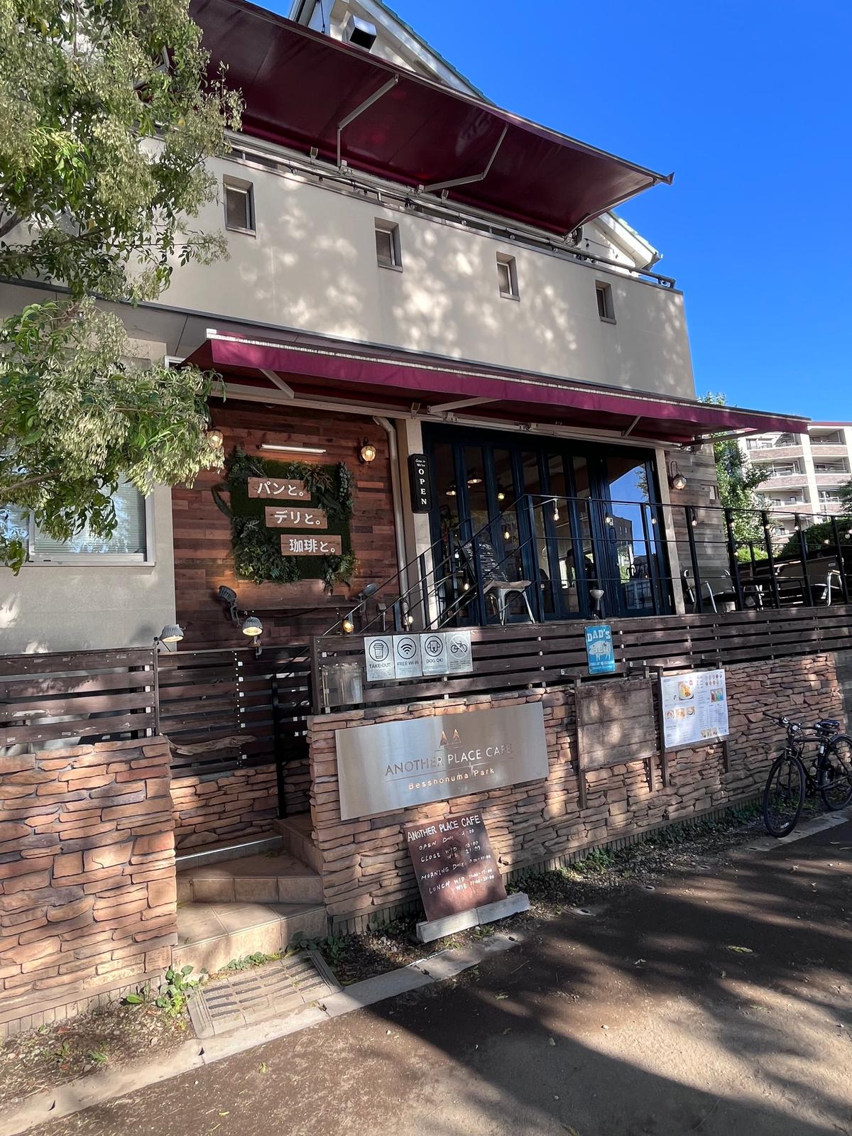 赤色の屋根とベージュ色の外壁のカフェ「ANOTHER PLACE CAFE」の建物。茶色のレンガの壁の前に黒い鉄柵があり、右側に自転車が停まっている。青空が背景。
