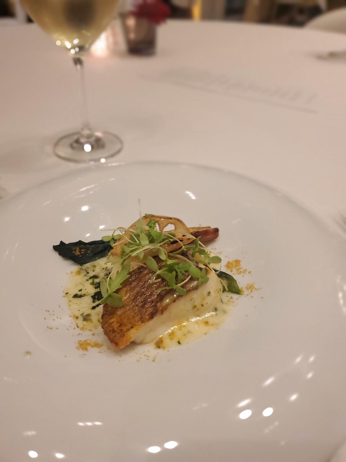 白磁の皿に盛り付けられた、淡いクリーム色のソースを添えた白身魚の料理。魚の表面は軽く焦げ目がついており、その上に緑色の葉と細切りになった玉ねぎ、マイクログリーンが散らされている。皿の縁には、黄色い粉末状のものがまばらに付いている。