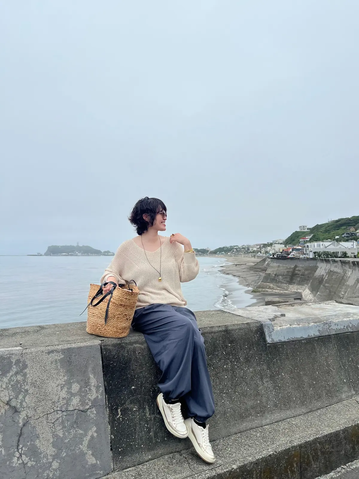 女性がコンクリート壁に腰かけて海を眺め、編みかごバッグを置いている。ベージュのセーターと紺色のズボン、ホワイトのスニーカーを履いている。海と遠くの街並みが見える。