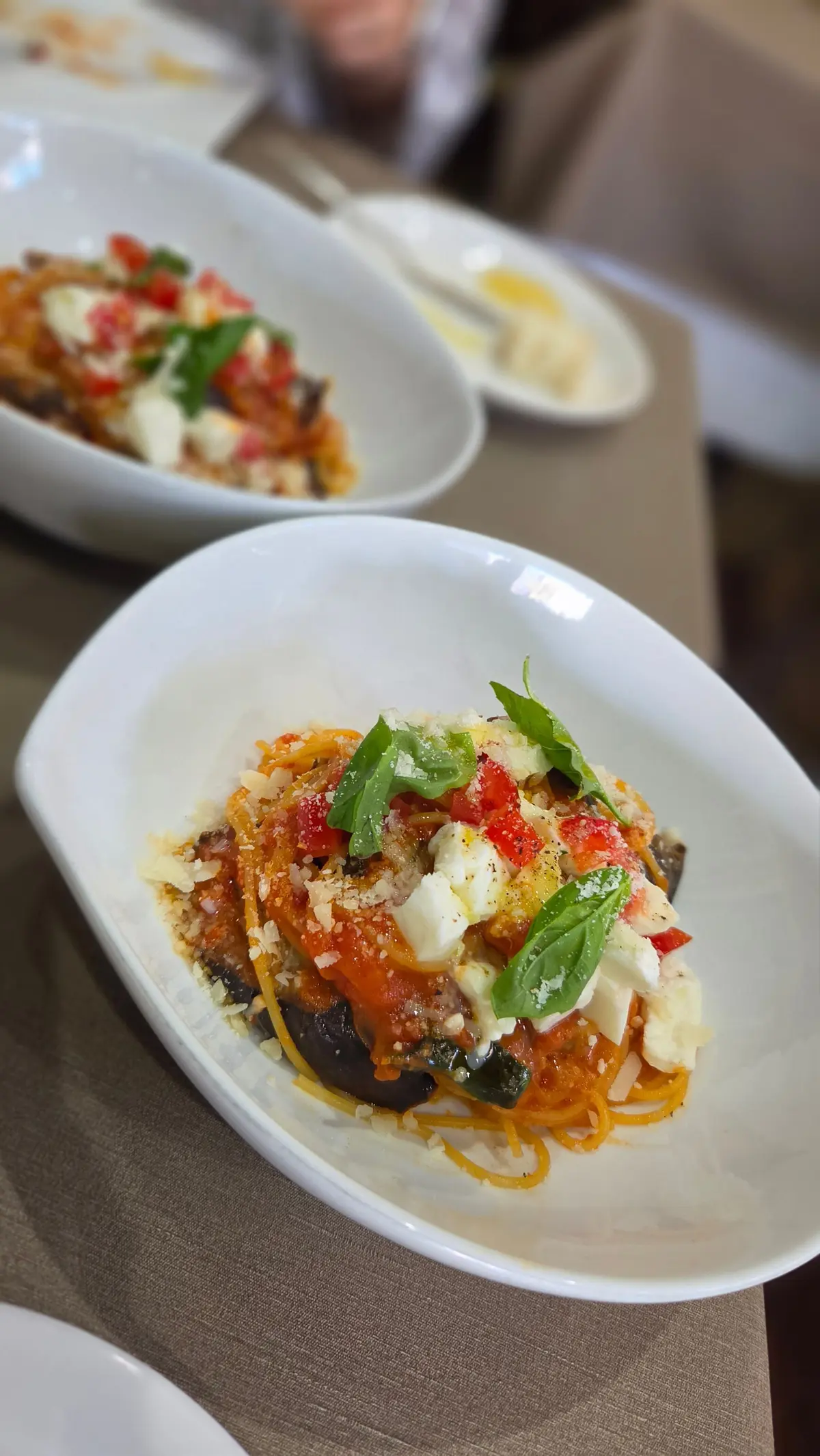 白い皿に盛り付けられたパスタ。トマトソースとチーズ、バジル、ナスがトッピング。背景には他の料理が写っている。