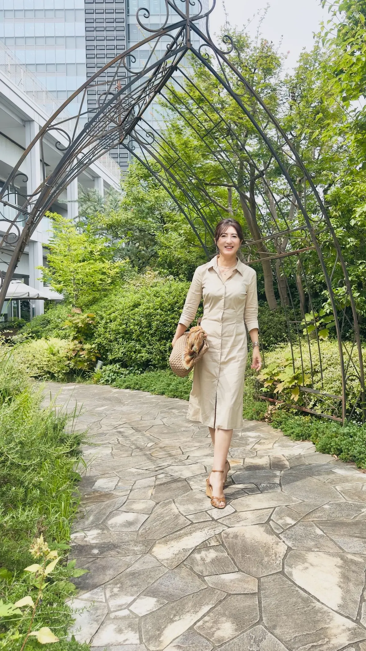 ベージュのシャツワンピースを着た女性が、石畳の道と黒い鉄製のアーチの下を歩いている。背景には緑豊かな植栽と高層ビルが見える。右手に編み込みのバッグを持っている。