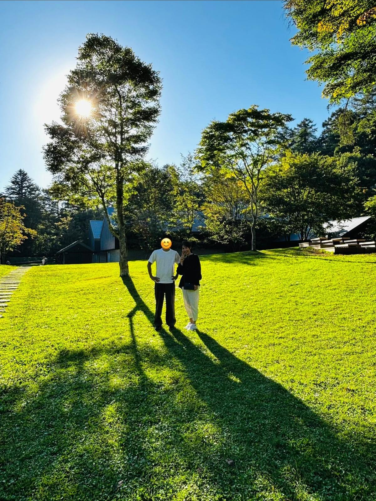 晴れた日の芝生の上、男女が寄り添って立っている。背後には木々が生い茂り、太陽の光が差し込んでいる。笑顔で顔を寄せ合う二人のシルエットが映える。