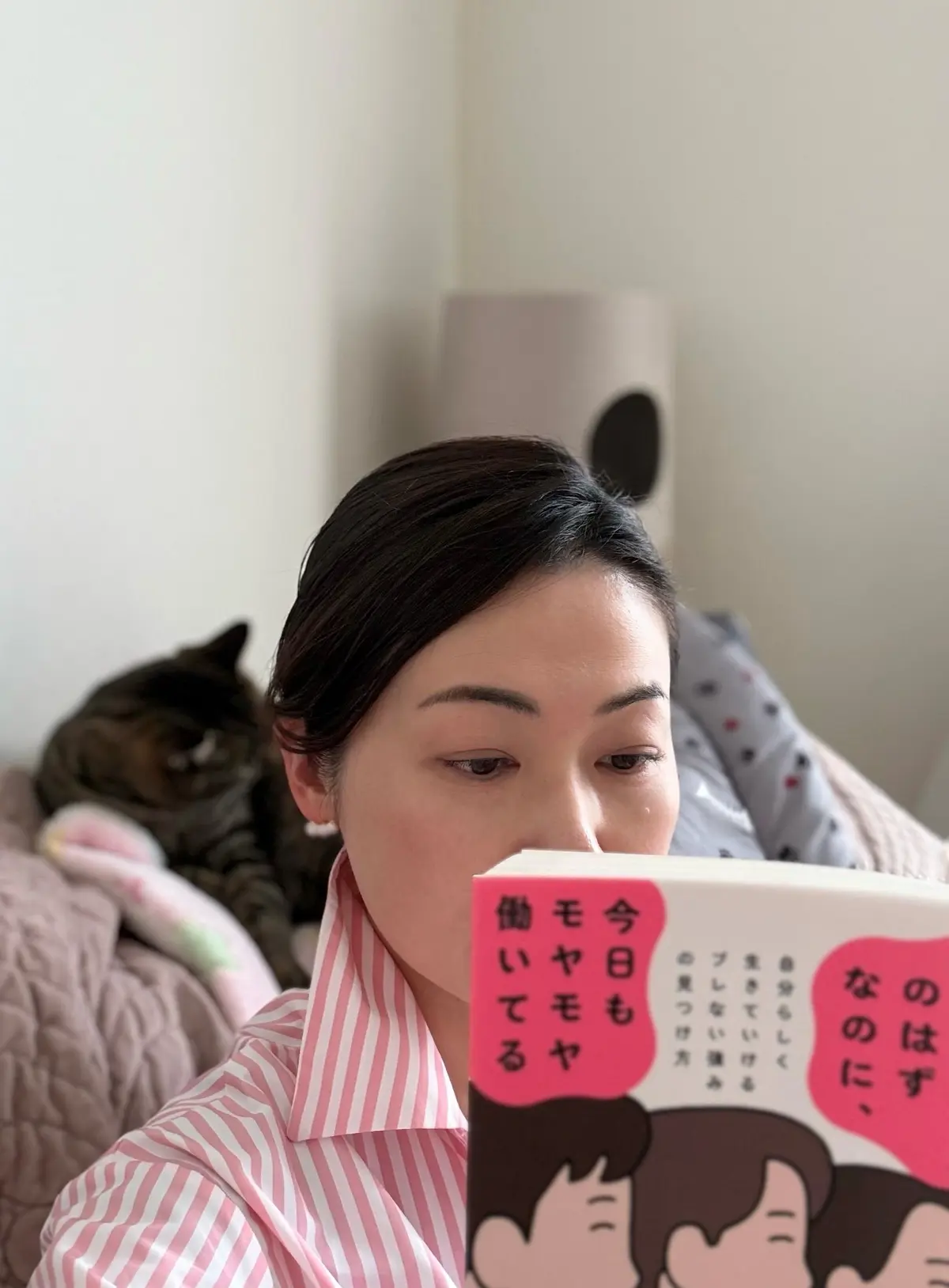 女性がピンクと白の縞模様のシャツを着て本を読み、猫が横に座っている