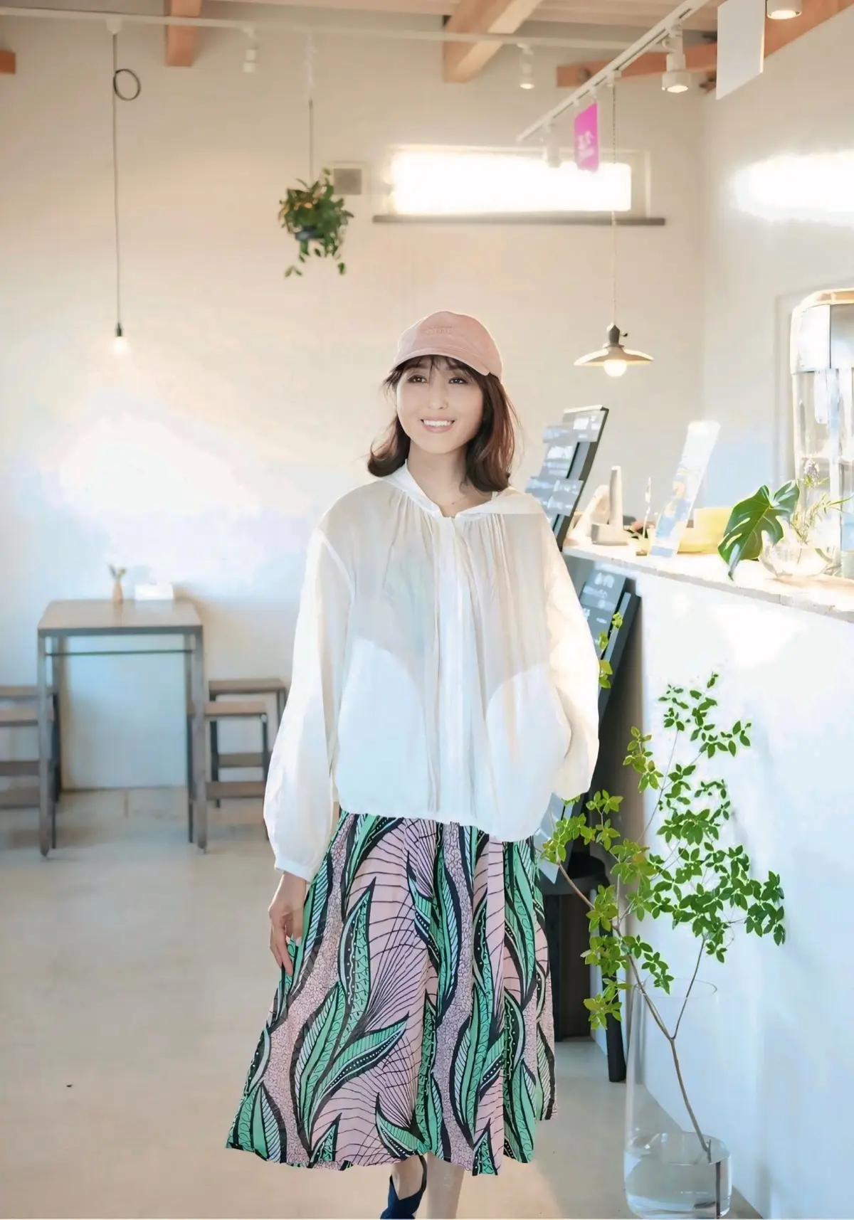 白いトップスに、ピンクと緑の柄のミモレ丈スカートを合わせた女性。ベージュのキャップを被り、右手にオフホワイトのバッグを持つ。木製の窓枠の前で微笑んでいる。