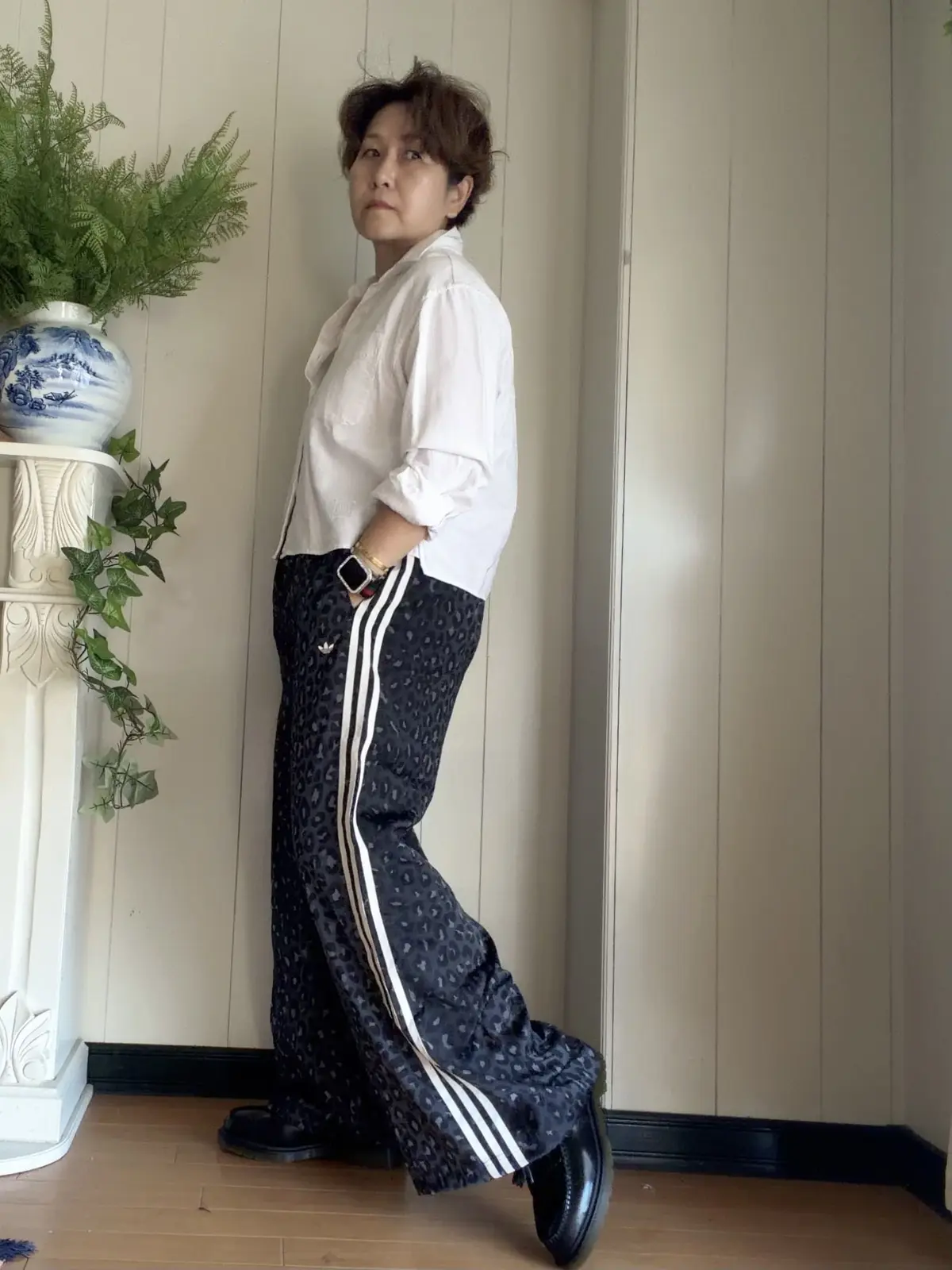 白いシャツにアディダスの柄パンツ、黒いブーツを履いた女性が立っている全身写真。背景には観葉植物と白い壁。