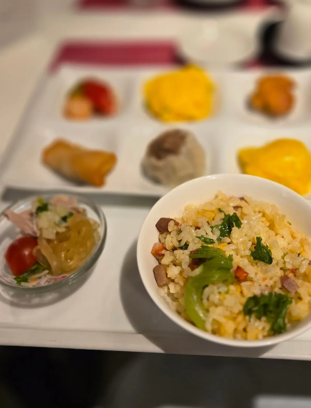 航空機内で提供される朝食。白いトレイに、チャーハン、点心、サラダ、パン、フルーツなどが盛り付けられている。背景には飲み物とカップが置かれている。