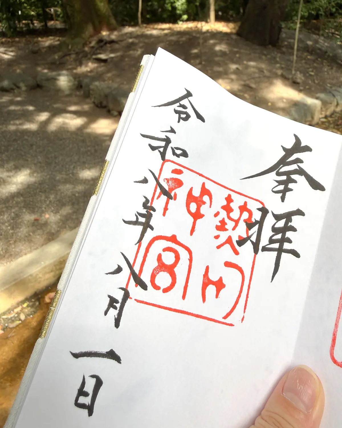 緑豊かな木々の背景に、手書きの日本語の書かれた紙。中央には赤い印鑑が押されており、日付が記されている。紙は開いており、指が添えられている。