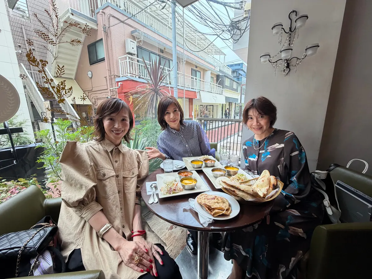 カフェのテーブルを囲み、笑顔の女性3人。手前にはインド料理と思われる料理が並び、背景に住宅街が広がる。