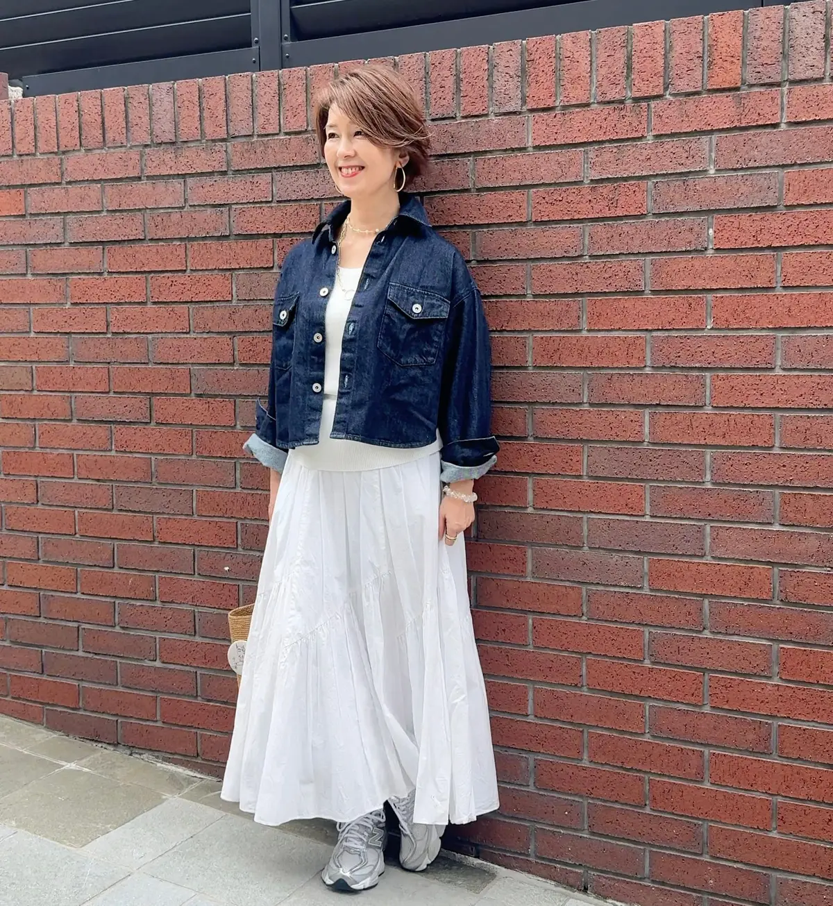 レンガの壁に寄り添って立つ女性。デニムジャケットと白いロングスカートを着て、笑顔でカメラを見つめている。足元にはグレーのスニーカーが履かれている。