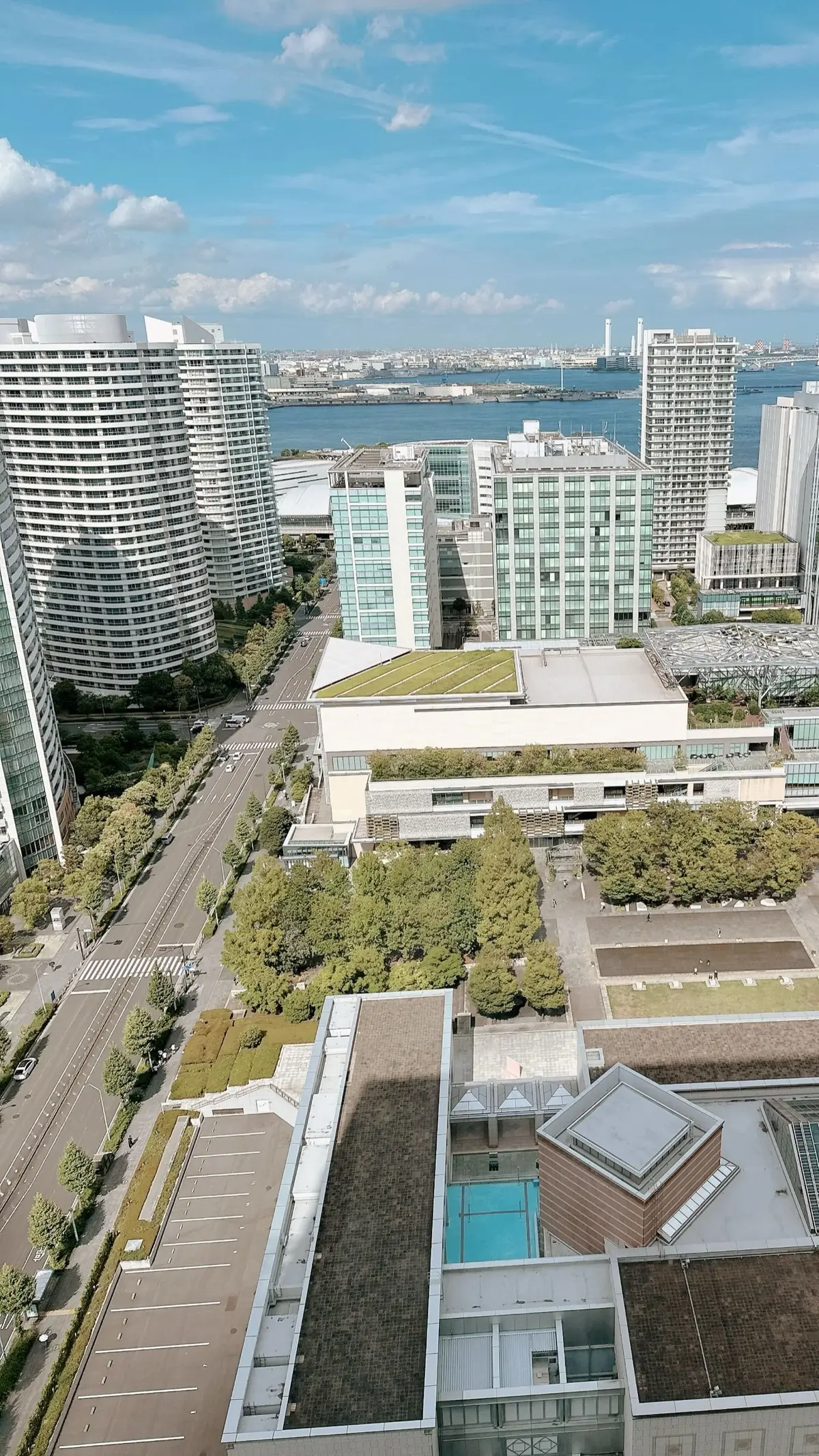 高層ビル群と港湾の風景。緑豊かな敷地に白い建物と水面が映るプール。青空を背景に、道路が通り、遠景に都市の建物が見える。