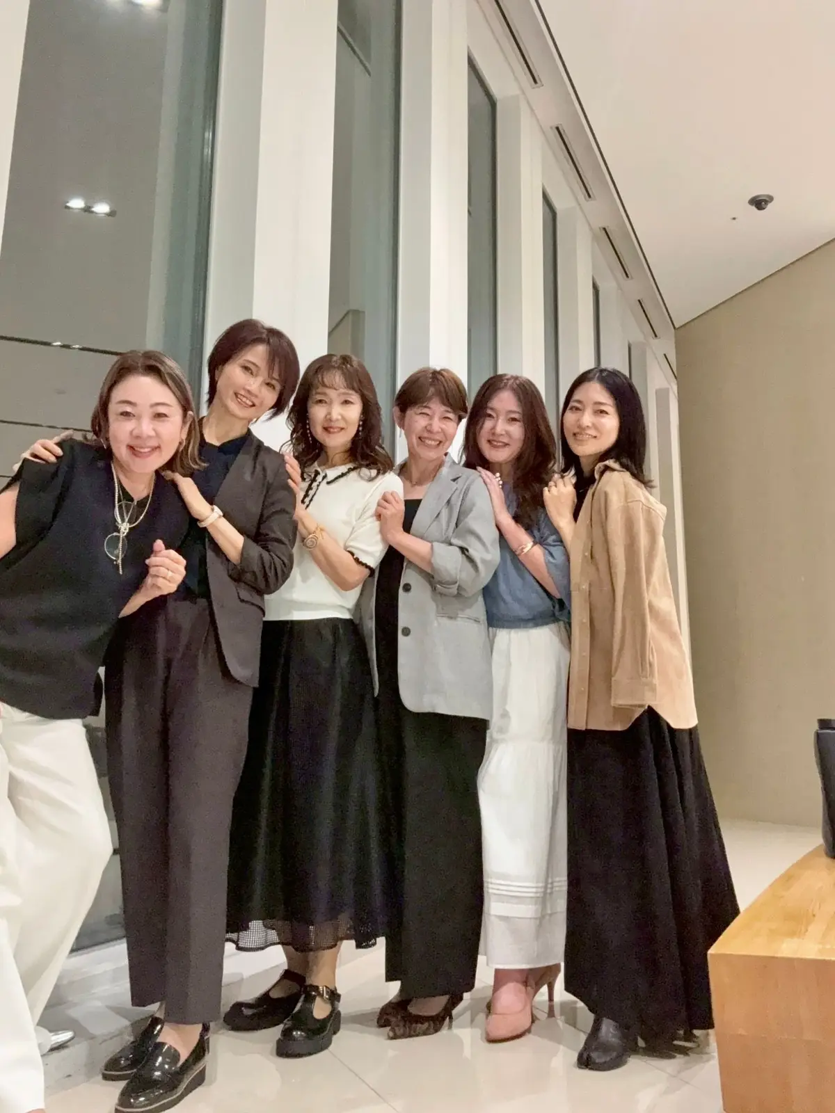 梅田ブルーバードでの誕生日ディナー女子会の集合写真 　6人の女性が明るく笑いながら、ガラスの壁のある建物の屋外で並んで立つ。左から順に、黒の服、ダークスーツ、白のトップス、グレーのカーディガン、ブルーのトップス、ベージュのカーディガンの服装。白い床に黒い靴を履いている。