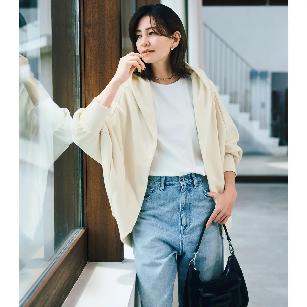 白いTシャツに淡い黄色のカーディガン、ブルーデニムを着用した女性。右手に触れ、横には黒いバッグを持つ。背景に木目調の窓枠と階段がある。