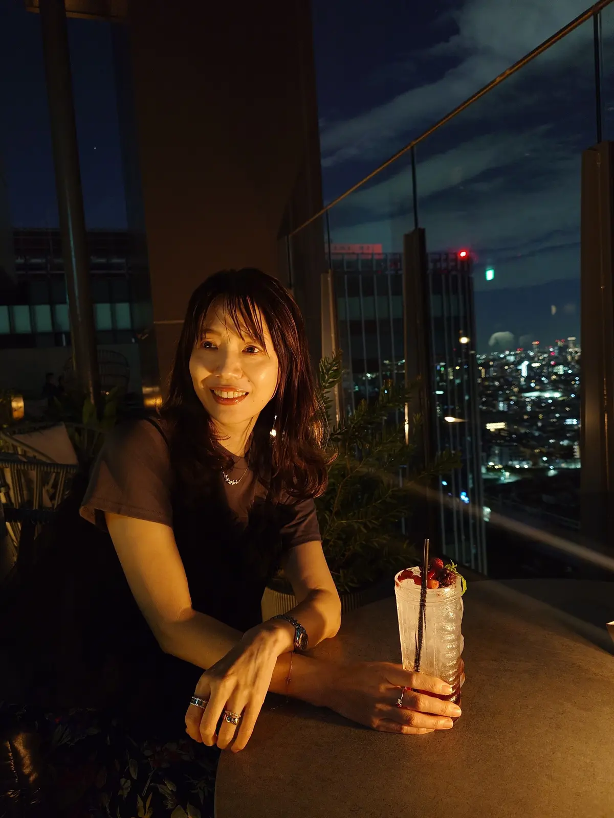 夜景を背景に、女性がテーブルに座ってくつろいでいる。黒いトップスに花柄のスカート。ジュエリーはペンダントがGRAFF、リングがCHANELとヴァンクリーフ&アーペル。手にはグラスに入ったドリンクを持ち、微笑んでいる。The TRAVELERS HOUSE ROOFTOP BARのルーフトップ。