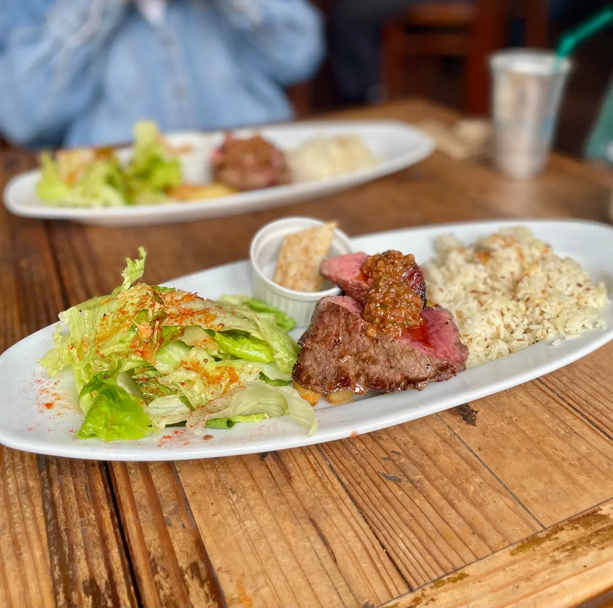 木製のテーブルに置かれた楕円形の白いお皿に、牛肉、レタス、ご飯が盛り付けられている。レタスには赤色の粉が振りかけられ、牛肉には濃い色のソースがかかっている。背景には、別の人が食事をしている様子が見える。