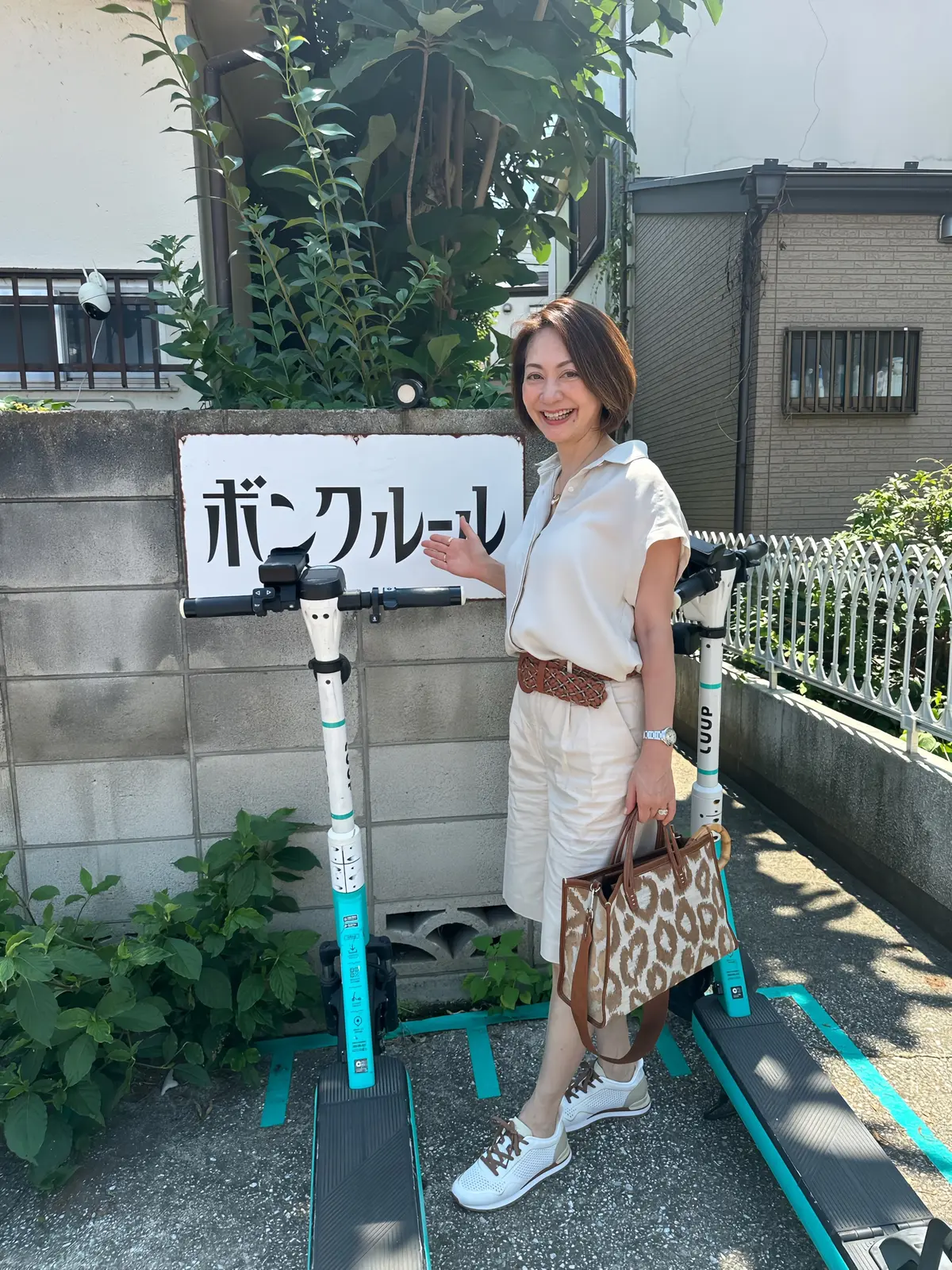 白いトップスに白いショートパンツ、白のスニーカーを履いた女性が、LOOOPの電動キックボードの横で微笑んでいる。背景には「ボンクール」と書かれた看板と緑の植物がある。