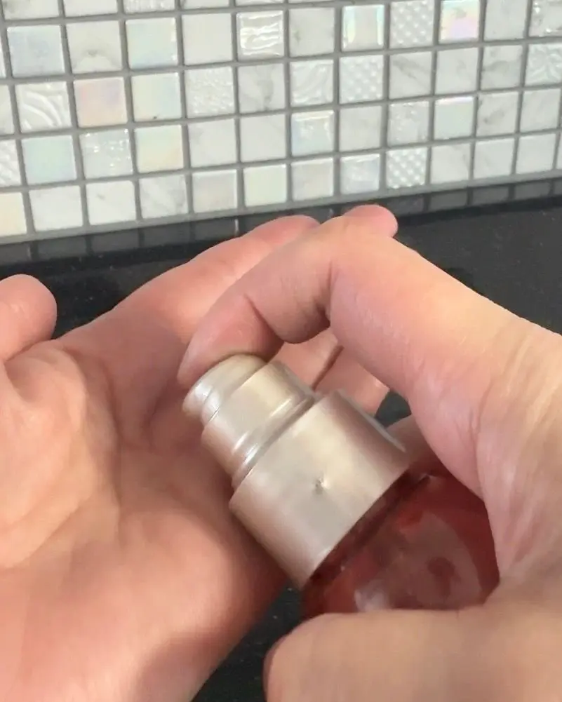 手のひらで支えられた、銀色のポンプ式の容器の蓋を指で押している様子。背景には白いタイルが貼られている。