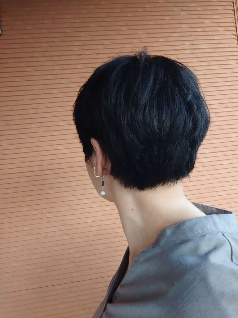 黒いショートヘアの背面が木製パネルの背景に映り、耳元には金具とパールが付いたイヤリングが輝いている