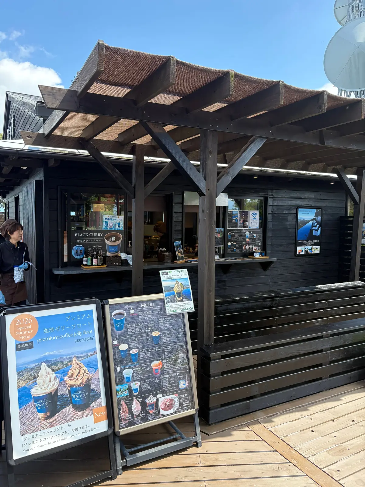 木造のフードスタンドとメニュー看板。屋根付きの建物には「BLACK CURRY」の看板が見え、右側にはメニューが掲示されている。手前にプレミアムコーヒーの看板が立っている。背景には青空とパラボラアンテナ。
