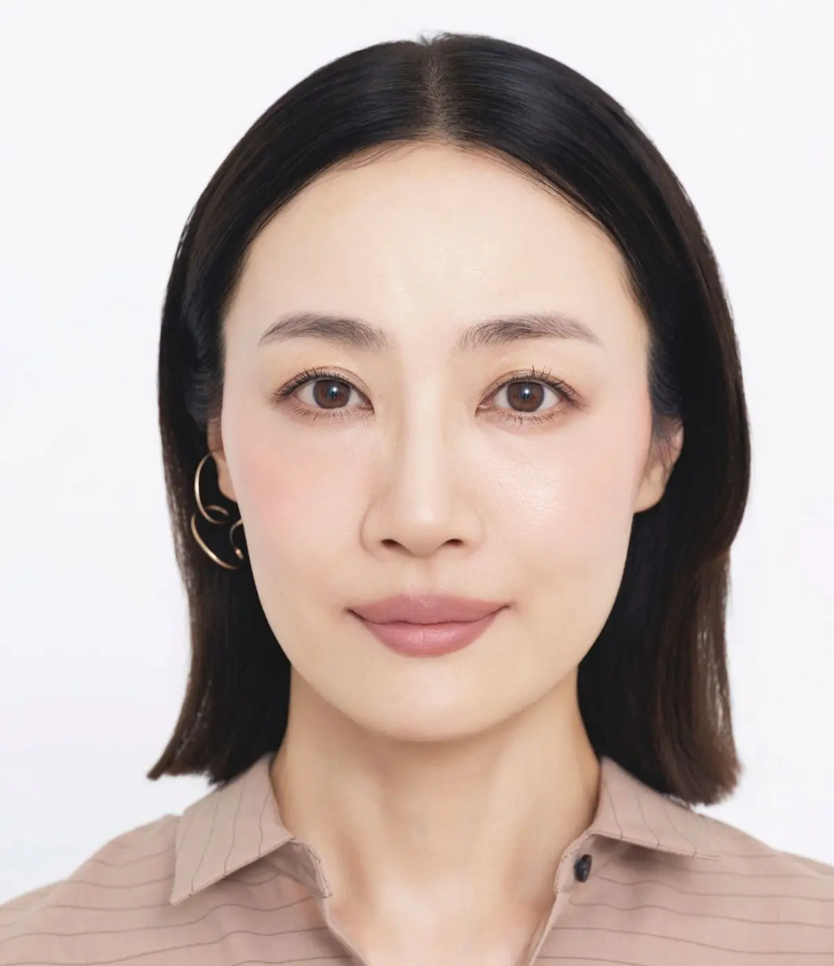 黒髪でショートカットの女性が、淡いピンクのストライプシャツを着て、ゴールドのイヤリングを装着している。白い背景に正面から撮影された。