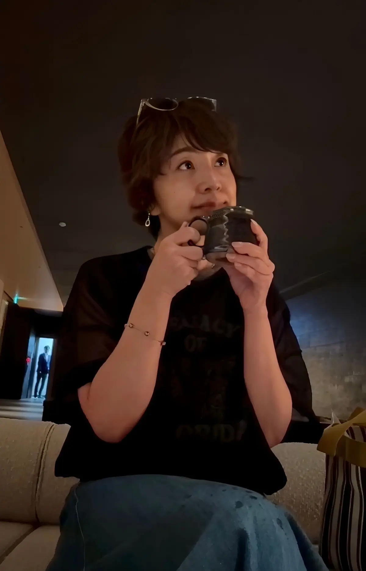 黒いTシャツを着た女性が、ガラス製のマグカップを持ち、こちらを見ている。茶色の短髪で、眼鏡をかけている。左腕にはブレスレット、右腕には腕時計をしている。背景にはソファと、遠くに人影が見える。
