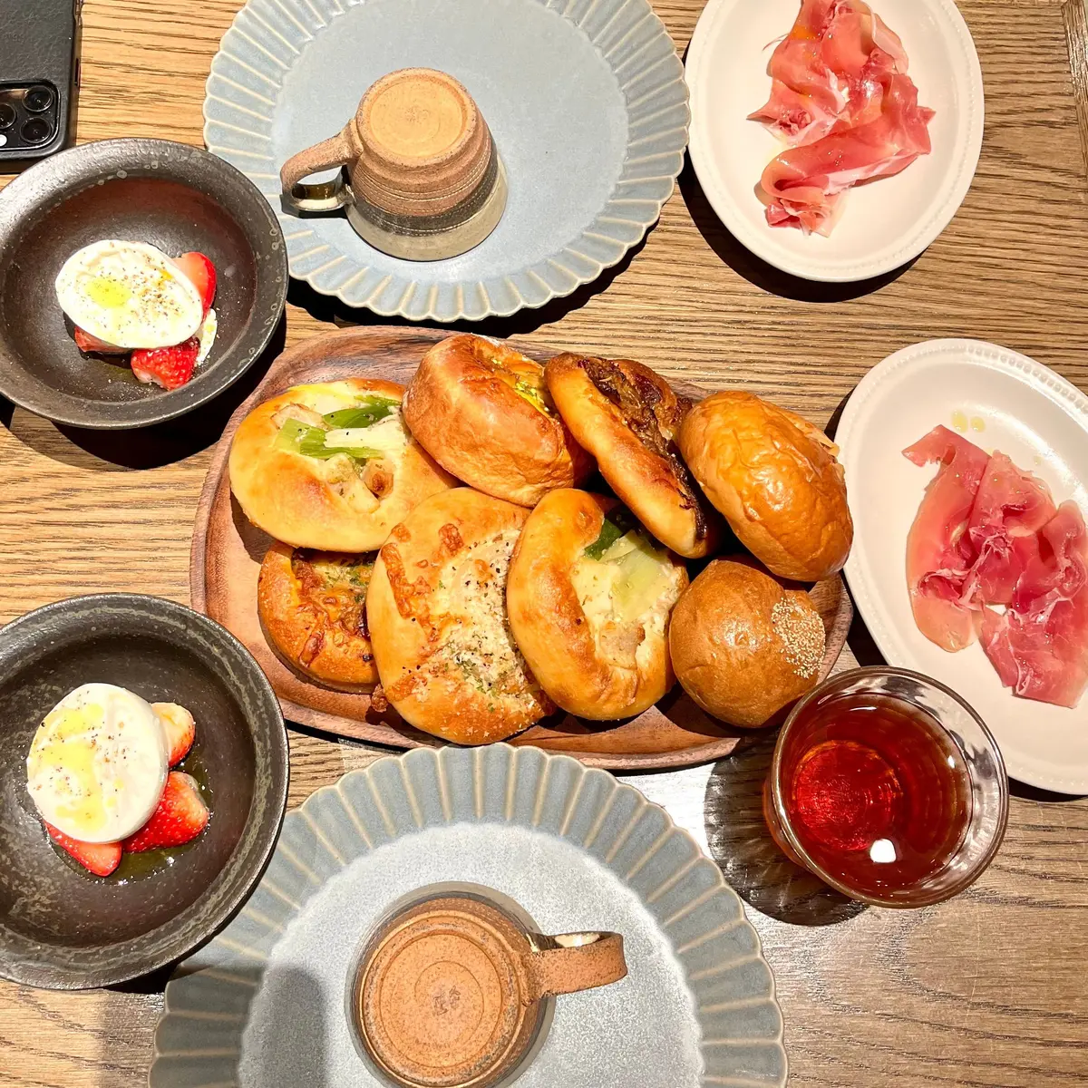 木製テーブルに、卵とイチゴの料理、焼きパンの盛り合わせ、薄切りハム、茶色のマグカップ、赤いドリンクが並んでいる