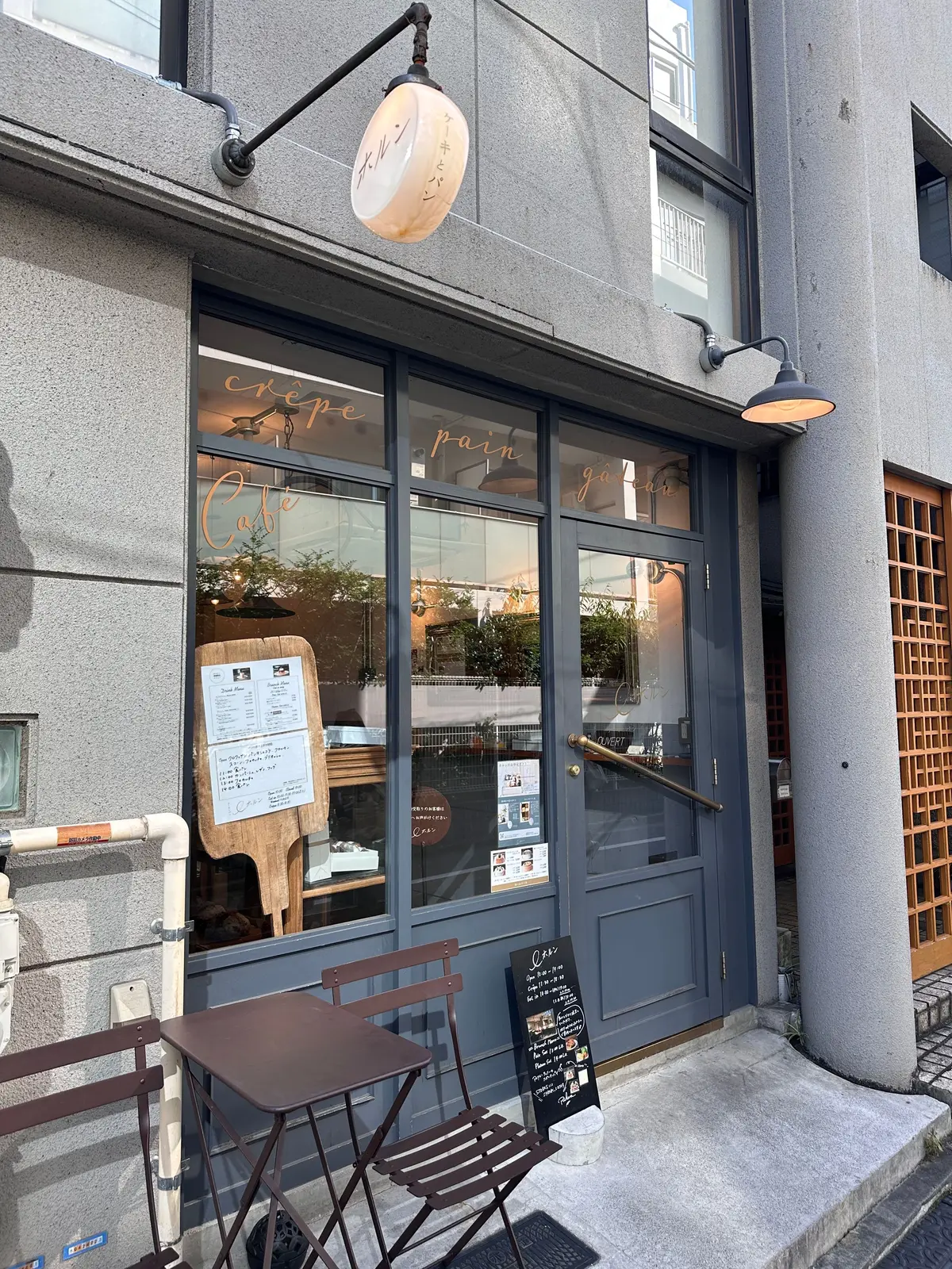 灰色の建物にある「crêpe pain gateau」と書かれたカフェの入り口。窓にメニューが貼られ、外にはテーブルと椅子が置かれている。右側には木製のドアがある。