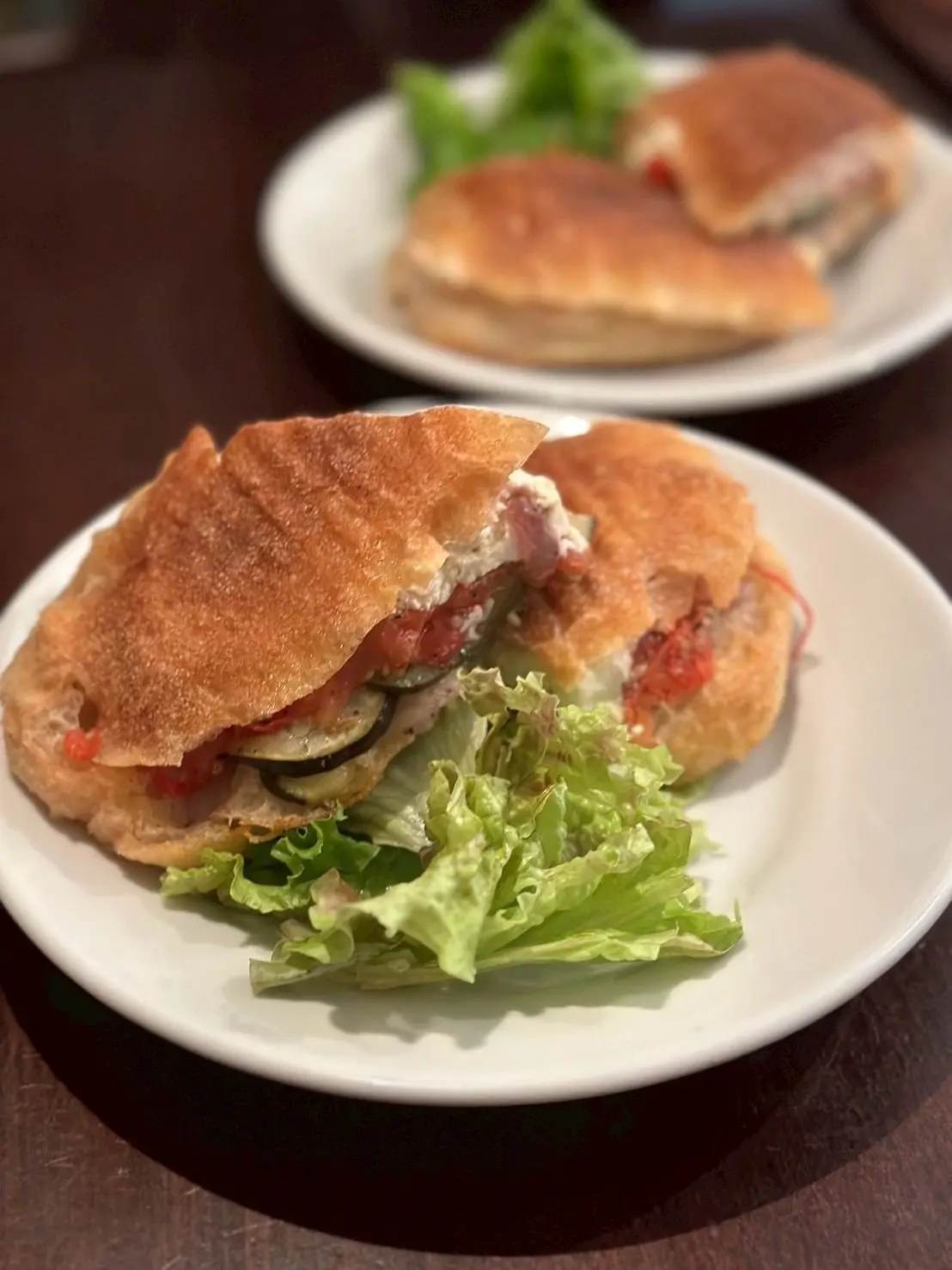 白磁の皿に盛り付けられた、焼き色のついたパン生地のパニーニ。中には、ズッキーニ、トマト、チーズ、レタスなどの具材が見える。背景には、同様のパニーニが別の皿に置かれている。