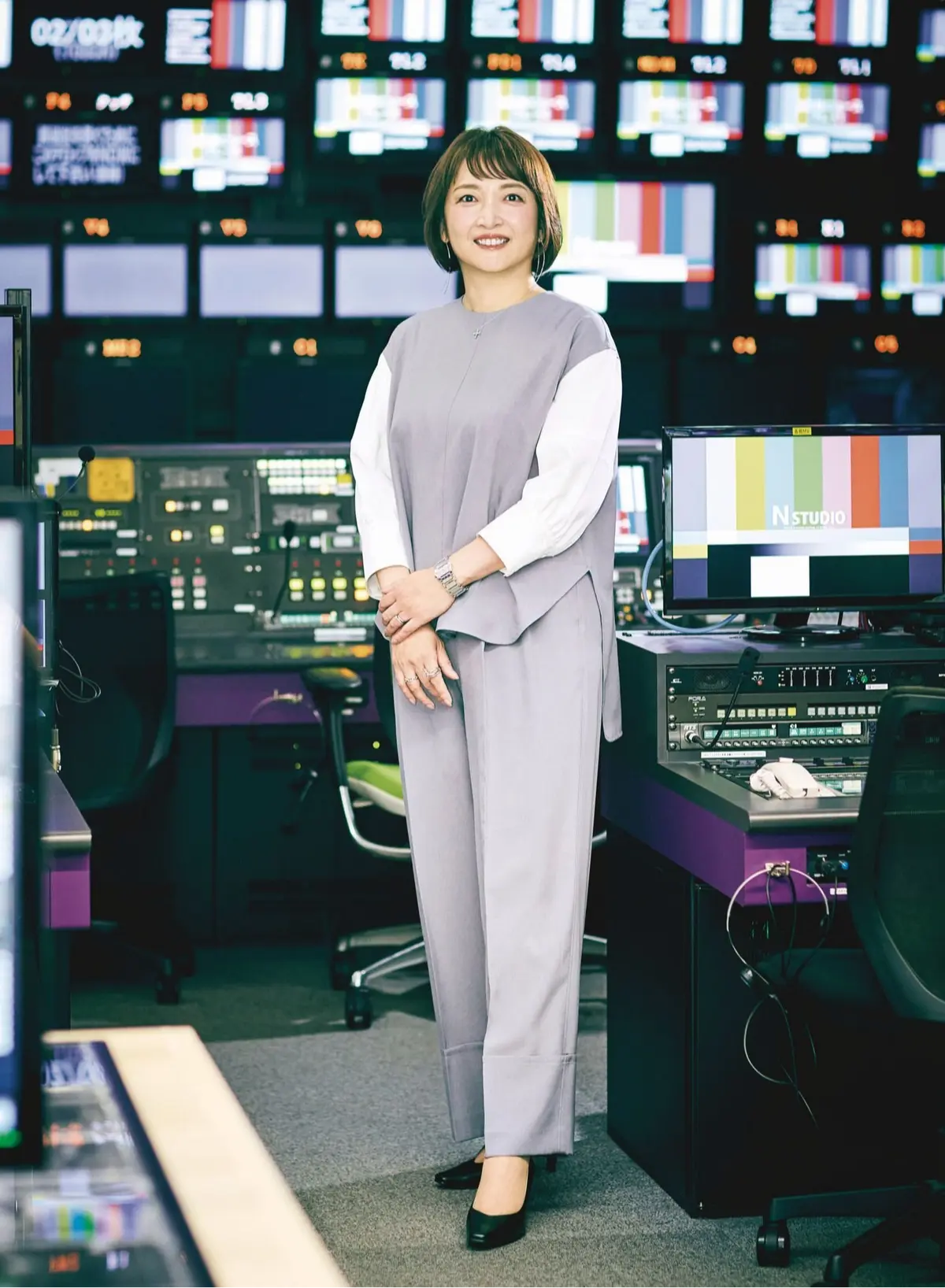 梅澤千佳子さん　エクラ華組　読者モデル　放送局の監視室で、グレーのトップスとパンツスーツを着た女性が微笑みながら立っている。背景には多数のモニターが並び、中央には「N STUDIO」と表示されたモニターがある。