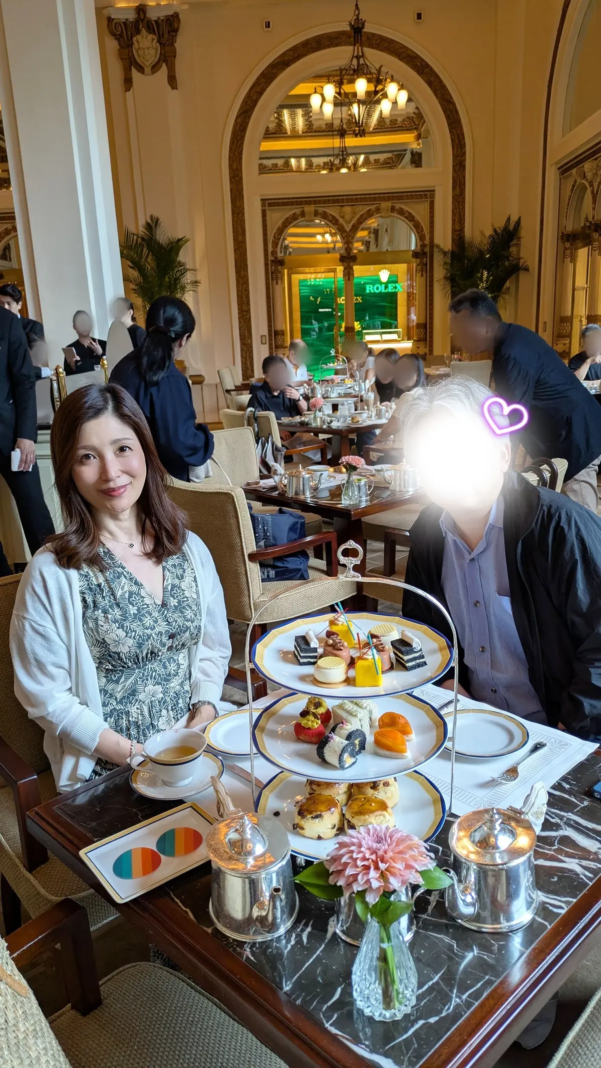 エレガントなカフェで、テーブルにはティーと多層のデザートプレート、銀のティーポット、花が置かれ、2人がリラックスして座っている