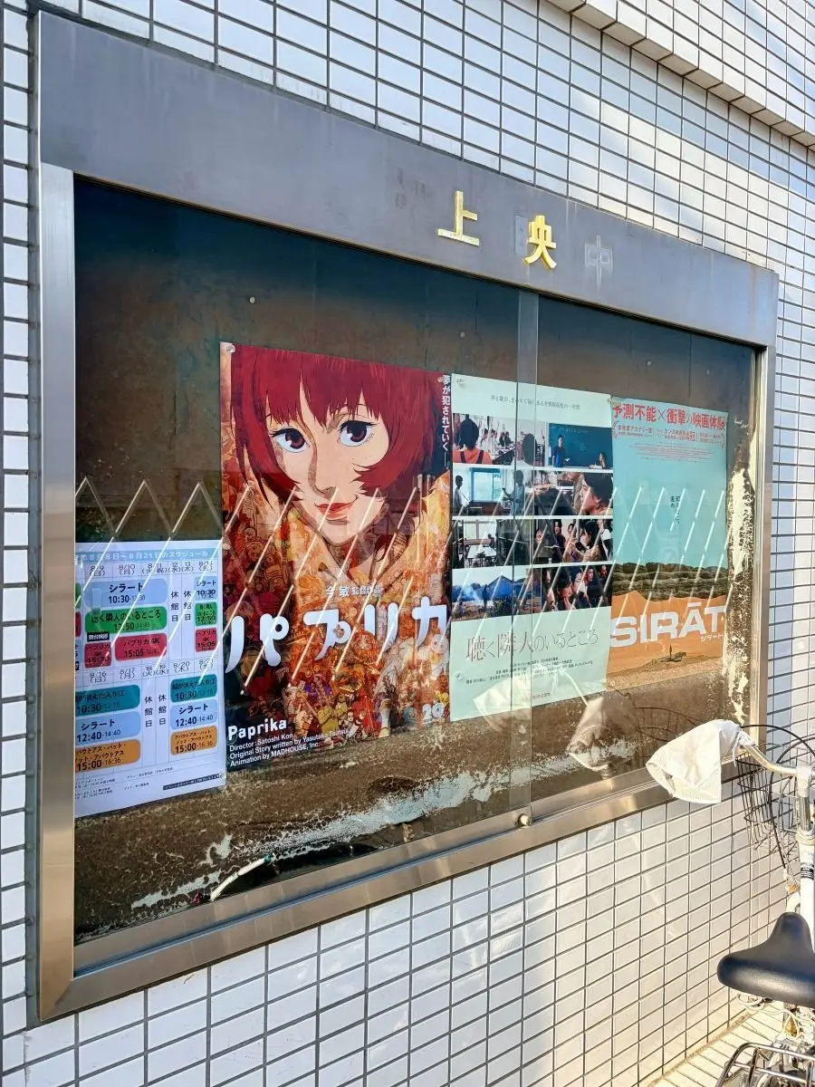 映画館の窓に貼られたポスター。中央には赤毛の女性が大きく描かれ、右隣には秋の風景が広がる「パプリカ」のポスター。窓の下には上映スケジュールが掲示され、自転車が置かれている。