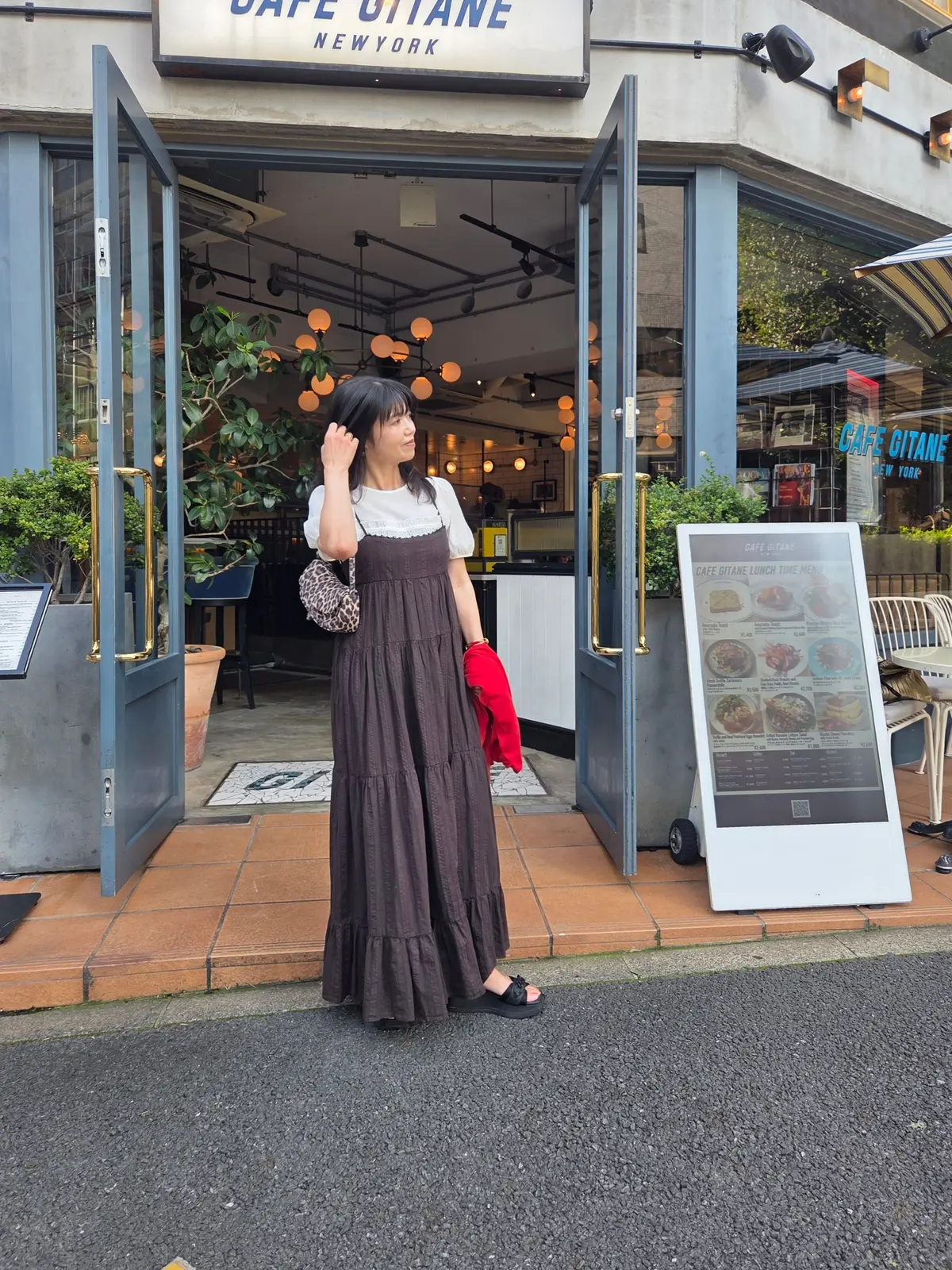 カフェ・ジタンの入り口に立つ女性。茶色のロングスカートに白いトップス、赤い手袋とレオパード柄のバッグを持つ。黒髪で、右手を上げてこちらを見ている。