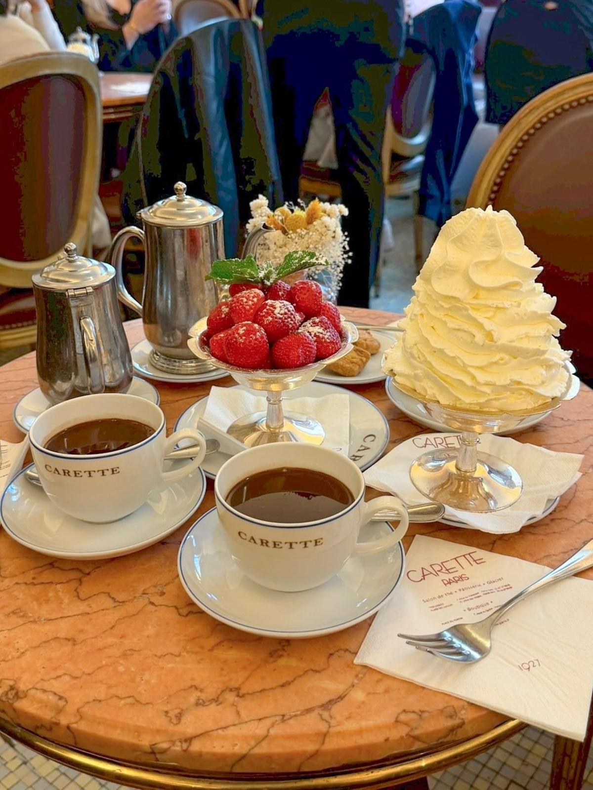 マーブル調のテーブルに、2つの「CARETE」ブランドのコーヒーカップ、銀製のティーポット、ストロベリーとクリームのデザート、フォークとメニューが置かれている