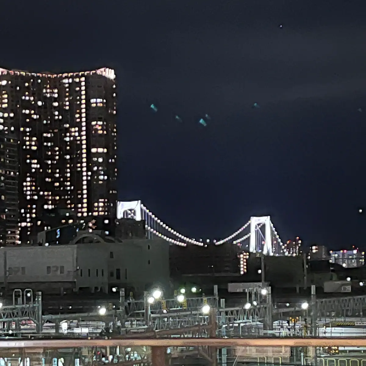 夜の東京の風景。高層ビル群と、ライトアップされたレインボーブリッジがそびえ立つ。手前には、工場のような建物と、電球が灯る構造物が見える。
