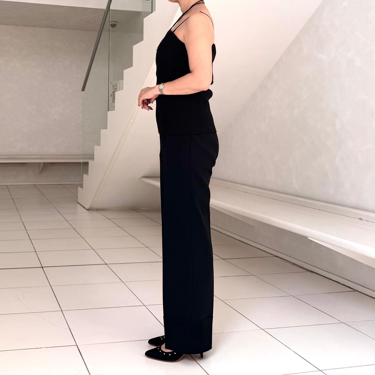 黒いニットタンクトップとパンツを着用した女性が、白いタイルの床に立っている横姿。右手に手を添え、左手首に腕時計をしている。背景には白い階段がある。