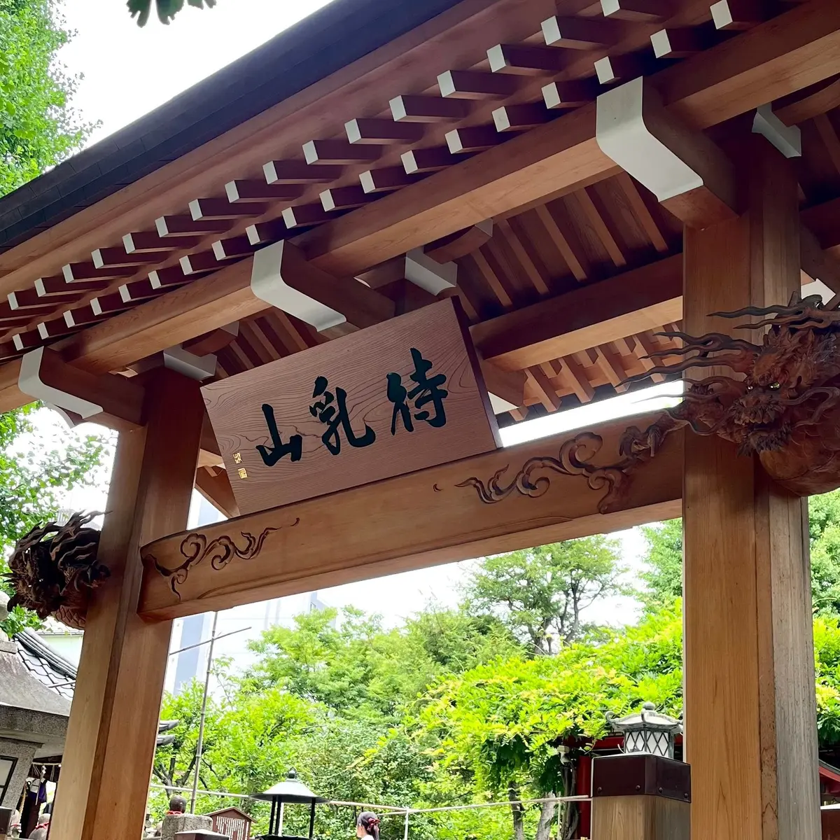 木造の柱と梁でできたアーチ型の門。中央には黒い文字で「山乳鉢」と書かれた木製の看板がはめ込まれている。背景には緑豊かな木々と、遠くに白い塔が見える。