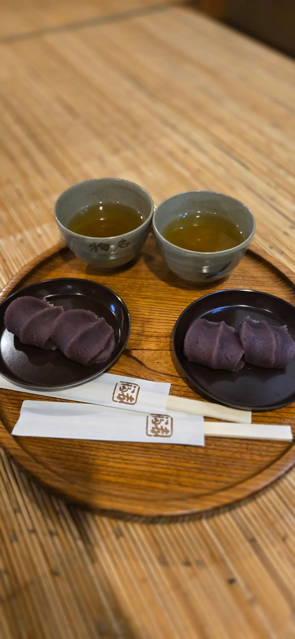 木製のトレイに、紫色の団子2個、お茶2杯、箸が置かれている。団子は小皿に盛り付けられ、お茶は灰色の湯呑みに注がれている。