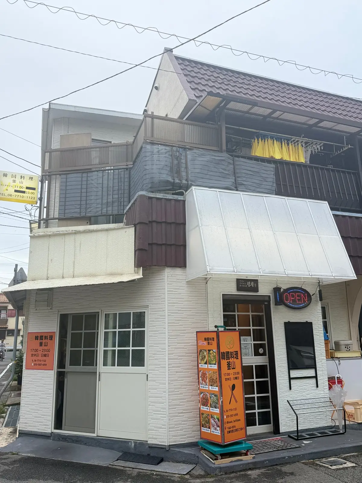 韓国料理 釜山 予約が取れないお店　尼崎グルメ　立花グルメ　パスポートのいらない韓国旅行　予約困難店　白いタイルの壁面を持つ二階建てのレストラン、入口にはガラスのドアと「OPEN」のネオンサイン、右側にメニュー表と看板が設置されている