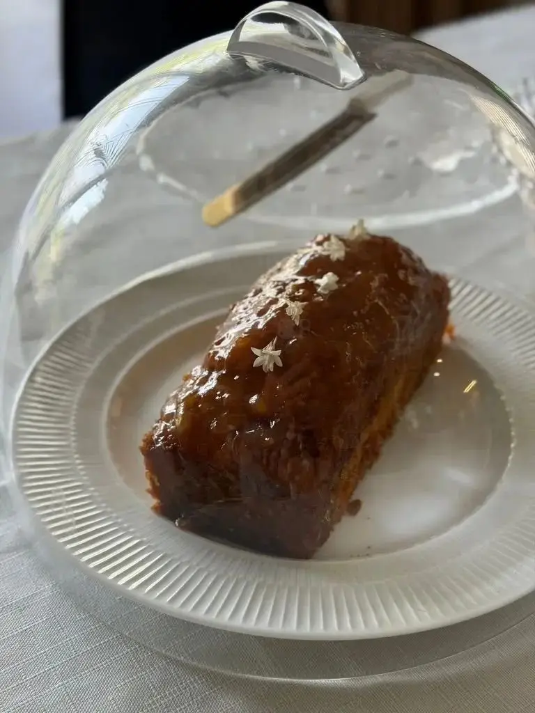 ガラスのドームが置かれた白いお皿の上、キャラメルソースをかけた四角い焼き菓子。表面には白い花びらと、削りたてのナッツが散らされている。