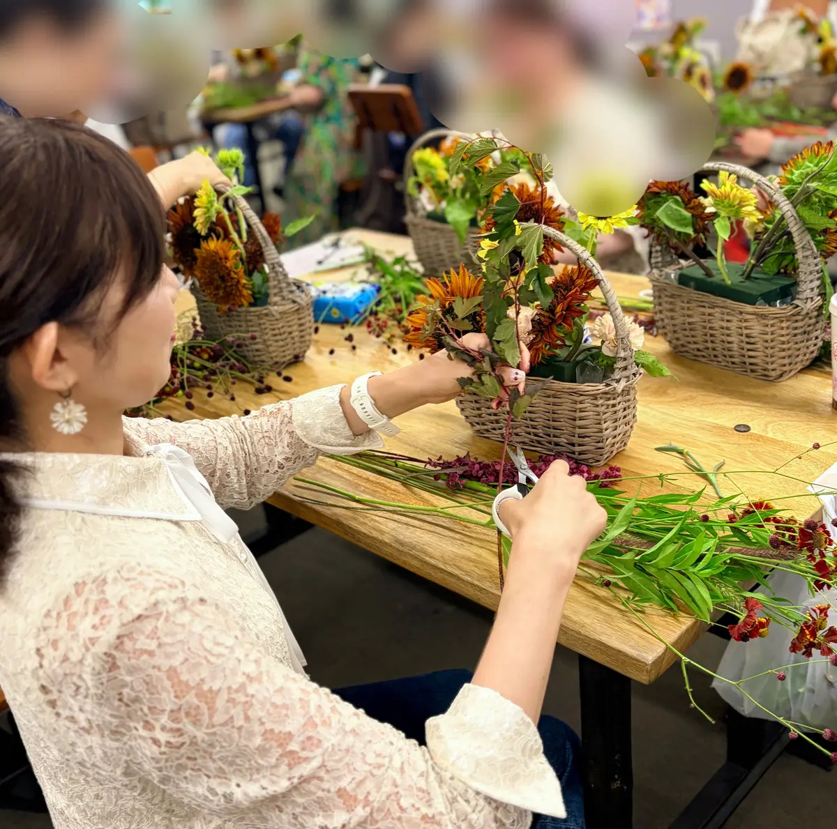 花束作り体験の教室。女性たちが編みのかごに、オレンジや黄色、赤色の花や葉を活けている。テーブルには複数の花材が並び、背景には植物やカラフルな布が見える。