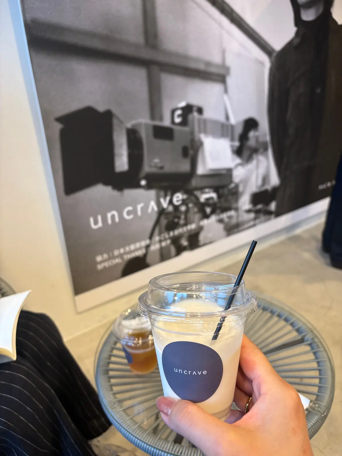グレーのテーブルに置かれた、Uncraveのロゴが入った白い紙カップに入ったアイスコーヒー。透明なプラスチックカップに入れ、黒いストローが挿さっている。背景にはUncraveのポスターが貼られている。