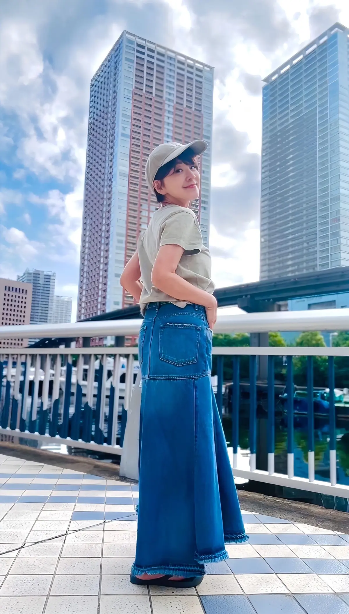 都会の背景をバックに、キャップを被った若い女性がポーズをとっている。ベージュのトップスに、裾がフリンジ状になった濃い青色のデニムスカートを着用。手は腰に当て、こちらを見ている。
