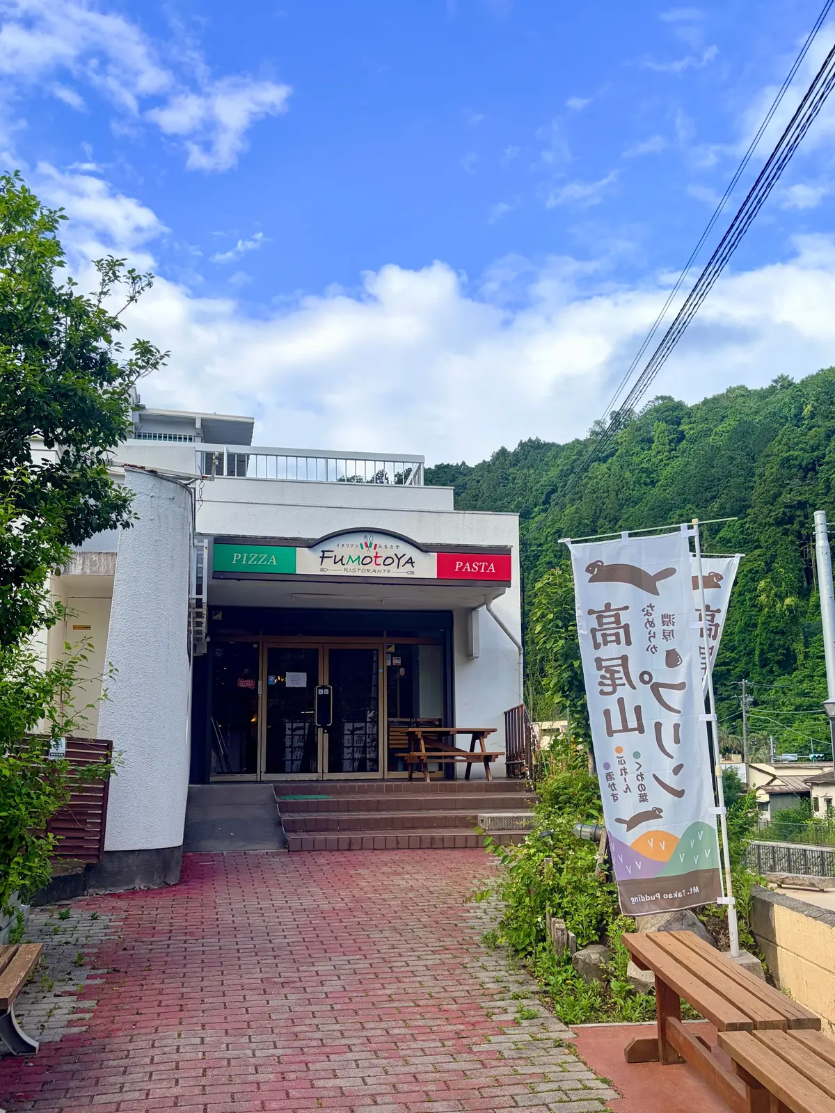 白い建物のレストラン「フモトヤ」の入口、緑の山と木々を背景に、赤いレンガの歩道とベンチが並ぶ
