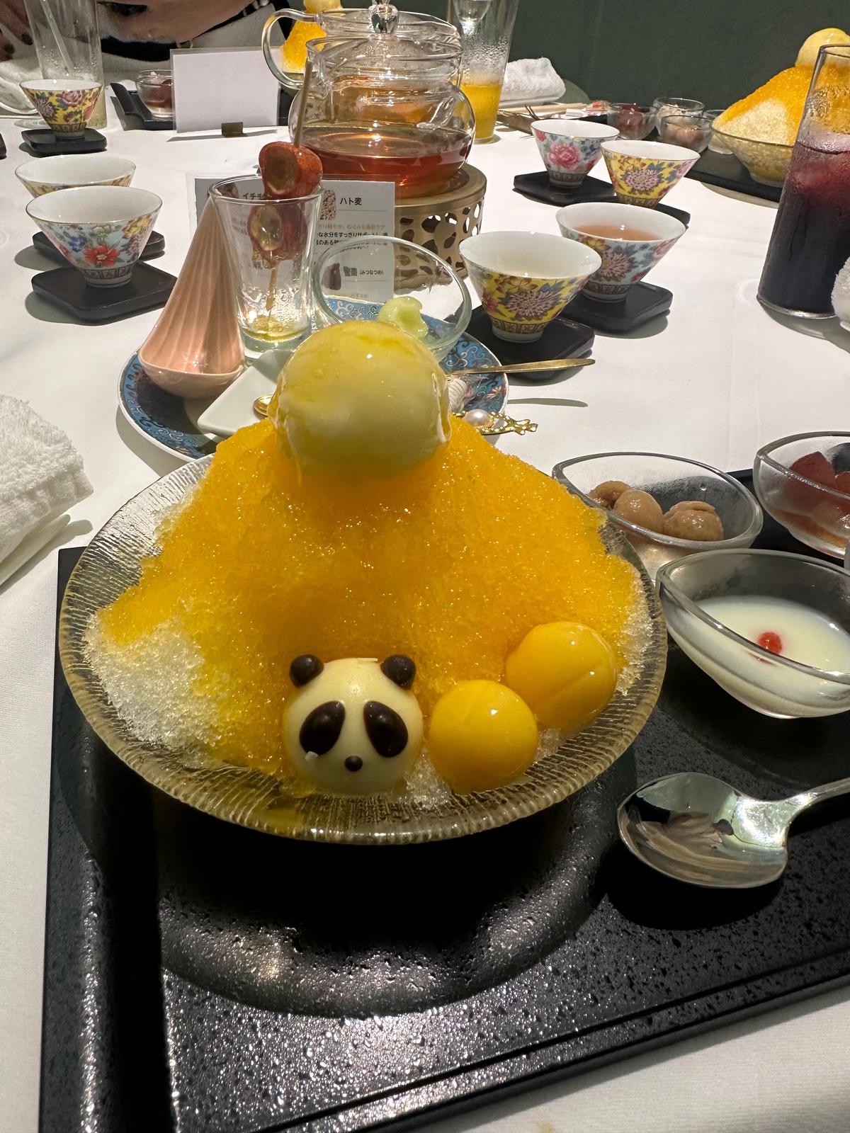 金色のかき氷に、パンダの形をしたゼリーと、黄緑色の丸いゼリーが添えられたデザート。黒いお皿に盛り付けられ、小皿に入った白玉や赤い豆菓子、飲み物と共に置かれている。