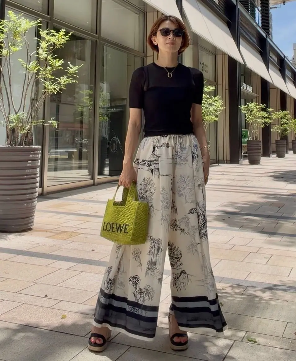 黒の半袖カットソーに、白黒の風景柄ワイドパンツを合わせた女性。緑色のLOEWEのバッグを持ち、ウェッジソールサンダルを履いている。背景にはガラス張りの建物と植栽がある。