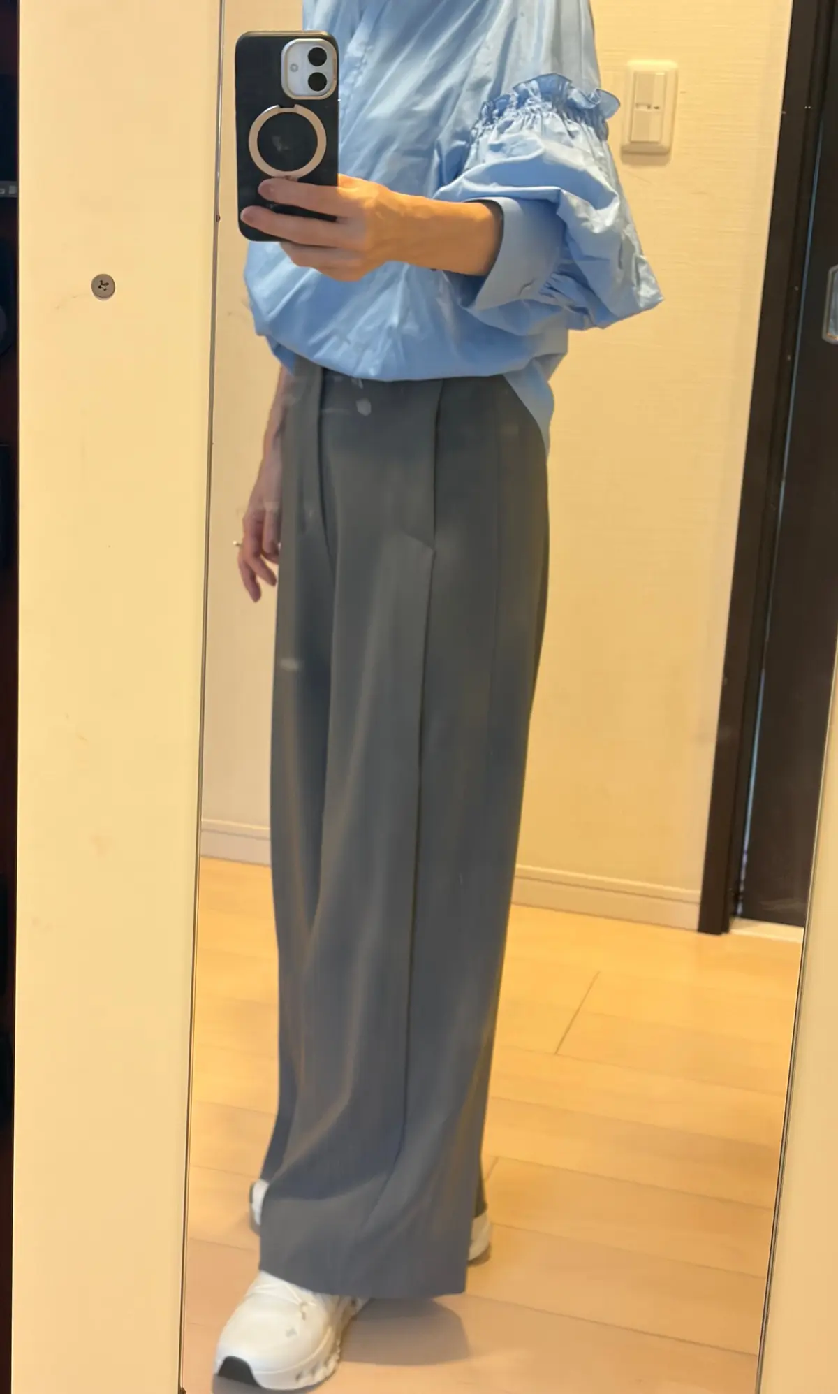 青色のパフスリーブシャツにグレーのワイドパンツを着用した人物の全身写真。白いスニーカーを履いている。背景にはドアと壁が見える。